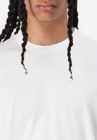 White crew-neck t-shirt with a smooth texture, featuring twisted hair strands on either side of a neutral face. Simple and unembellished design.