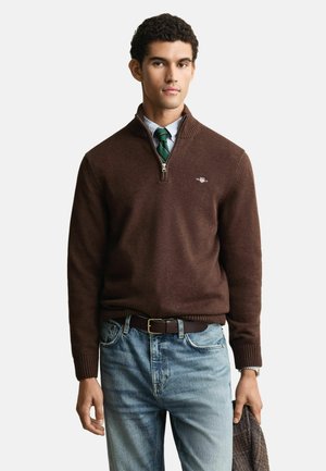 Brauner Zip-Hals-Pullover aus weichem Material, mit gerippten Bündchen und Saum. Kombiniert mit hellblauen Jeans und einem dunklen Gürtel.