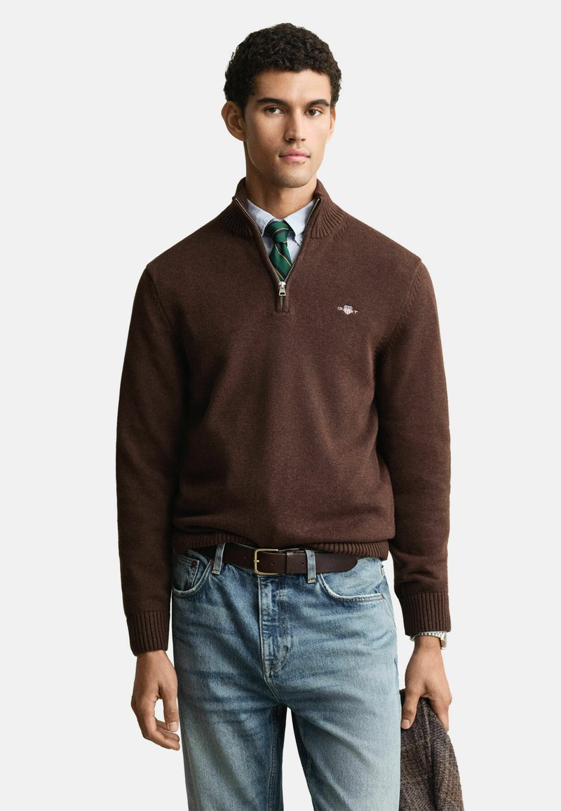 Brauner Zip-Hals-Pullover aus weichem Material, mit gerippten Bündchen und Saum. Kombiniert mit hellblauen Jeans und einem dunklen Gürtel.