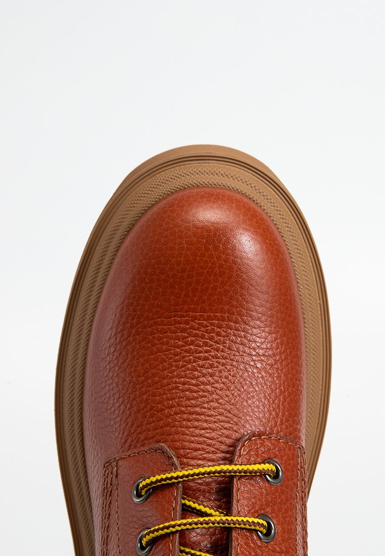 Brown leather shoe with textured finish, round toe, and yellow laces. Features a thick tan sole with ridged pattern.