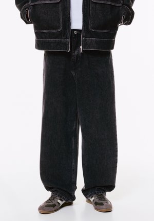 Persona che indossa pantaloni di jeans neri larghi, giacca di jeans nera, camicia bianca e sneakers grigie con accenti scuri e bianchi.