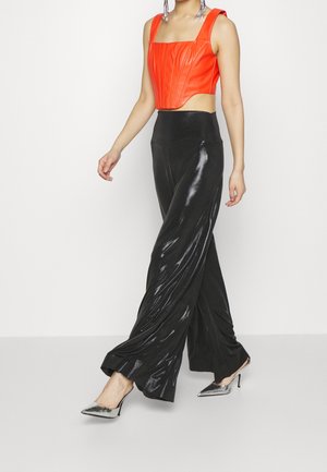 Femme marchant avec un pantalon large noir brillant, un corset sans manches orange vif, des chaussures à talons hauts argentées et de longues boucles d'oreilles pendantes.