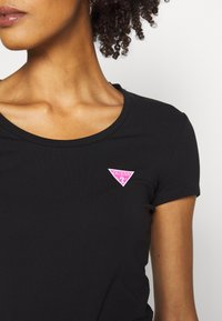 Zwarte getailleerde t-shirt met korte mouwen en een ronde halslijn, voorzien van een roze driehoekig logo met het woord "GUESS" op de linkerborst.