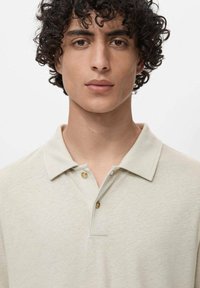 Polo de tela beige claro, con tapeta de botones y una textura suave. Cuello estándar con un corte relajado. Sin patrones ni acentos.