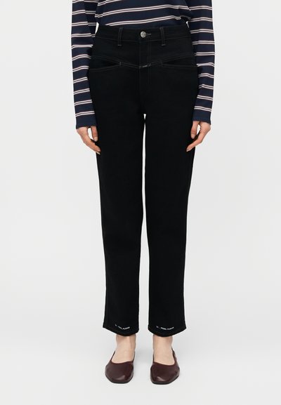 Jean noir à coupe droite avec poches avant et arrière, porté avec un haut à manches longues rayé et des ballerines rouge foncé, posé devant un fond blanc.