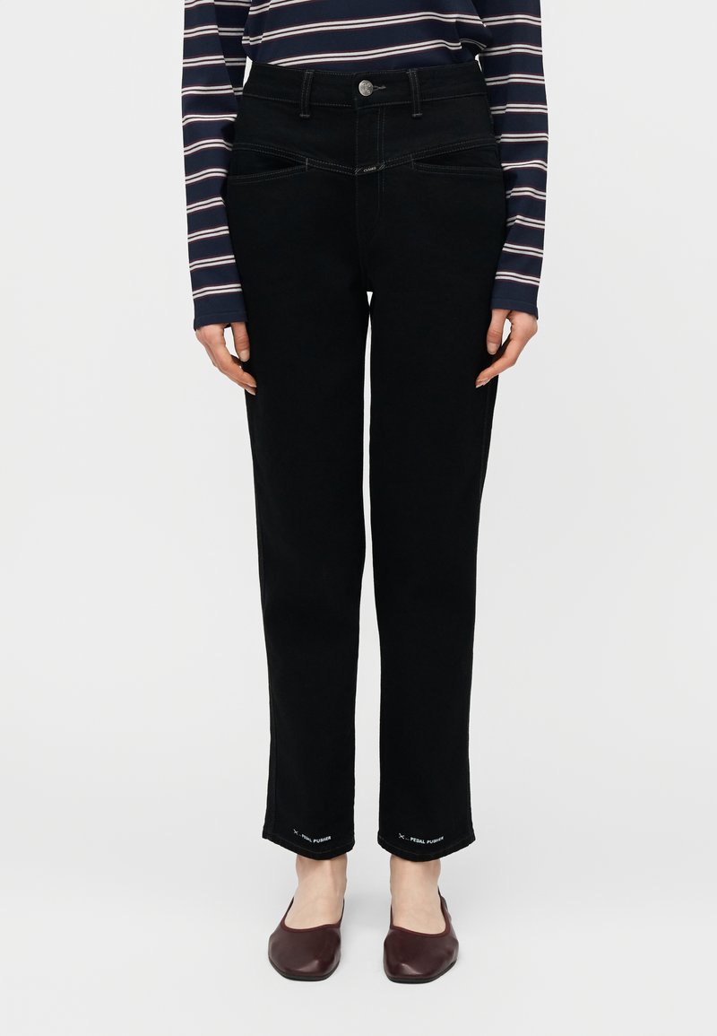 Μαύρο τζιν με ίσια γραμμή και τσέπες μπροστά και πίσω, φορεμένο με ριγέ μακρυμάνικη μπλούζα και σκούρα κόκκινα μπαλαρίνες, στέκεται μπροστά σε λευκό φόντο.