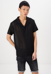 Black short-sleeve shirt in lace material, featuring a collar, button closure, and textured pattern with vertical lines and openings.