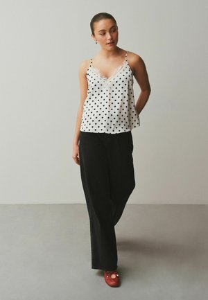 Žena stojaca pri svetlej stene, oblečená v bielom tielku s bodkami, čiernych nohaviciach s širokými nohavicami a červených baletných topánkach, pozerajúca sa do strany.