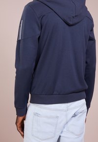 Marinblå hoodie med texturerad huva, ribbade ärmslut och nederkant. Har vita accentrandar på högra ärmen och en slät tygfinish.