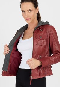 Rote Lederjacke mit grauem Kapuzenbesatz, ausgestattet mit einem Reißverschluss vorne, seitlichen Taschen und einem weichen Innenfutter. Glatte Textur, taillierte Passform.