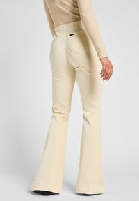 Persona che indossa pantaloni a zampa d'elefante in corduroy a coste color crema e una maglietta beige a maniche lunghe, vista da dietro su uno sfondo neutro.