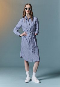 Rutet skjortekjole i blått og hvitt, med belte i midjen, lange ermer, knappeåpning foran, og kombinert med hvite sneakers og sokker.