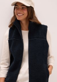 Námořnický fleecový vest s vysokým límcem, zipovým uzávěrem a bočními kapsami přes lehký svetr. Hebká textura a útulný design pro vrstvení.