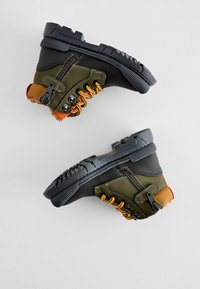 Paire de chaussures de randonnée noires et vert olive avec lacets orange, fermetures éclair latérales et semelles noires robustes sur fond clair.