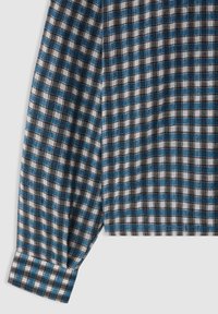 Cuff e parte della manica di una camicia a maniche lunghe a scacchi blu, nera e bianca su uno sfondo grigio chiaro.