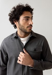 Cămașă gri cu buton pe care se află un buzunar pe piept, mâneci lungi și o textură netedă; stratificată peste un tricou alb. Modelul își ajustează gulerul cu mâna.