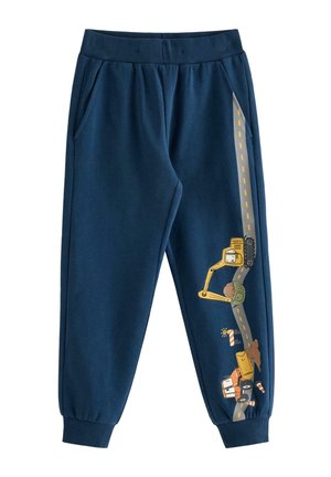 Lächelndes Kind mit lockigem blondem Haar, das einen blauen Pullover und eine Hose mit Baustellenfahrzeug- und Straßendesigns trägt, steht vor weißem Hintergrund.