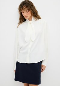 Blouse blanche avec un col à nœud, manches longues et coupe légèrement ample, associée à une jupe marine. Tissu lisse et léger.