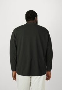 Černé tričko s dlouhým rukávem, volnějšího střihu, kulatým výstřihem a hladkou látkou; pohled zezadu ukazuje švy na ramenou a rovný spodní lem.