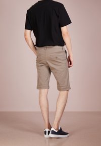 Shorts kaki avec ourlets retroussés, deux poches arrière avec accents de bouton, portés avec un t-shirt noir et des chaussures noires décontractées. Texture de tissu lisse.