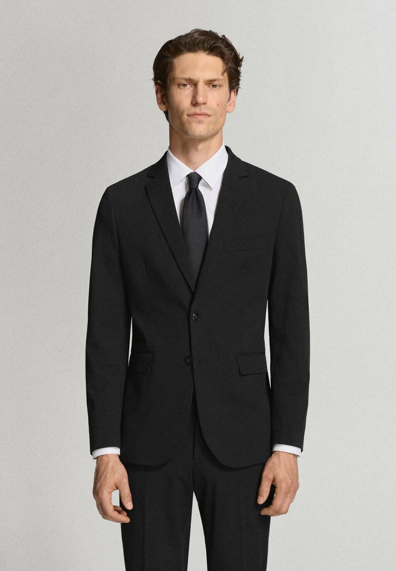 Homme portant un costume noir cintré avec une chemise blanche et une cravate noire, se tenant devant un fond gris clair uni.