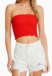 Top corto rojo sin tirantes con un patrón texturizado y ondulado, y dobladillo con volantes, combinado con shorts de mezclilla blancos desgastados con bordes deshilachados.