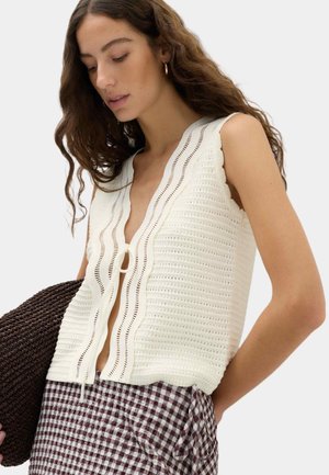 Frau mit langen, welligen Haaren, die ein weißes ärmelloses Stricktop mit Bindeband vorne und karierte braune und weiße Hose trägt und eine dunkel gewebte Tasche hält.