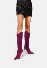 Stivali alti in suede viola con punta affusolata e tacco a stiletto. Presentano una morbida texture e un design con risvolto, che si abbinano a una gonna in pelle nera.