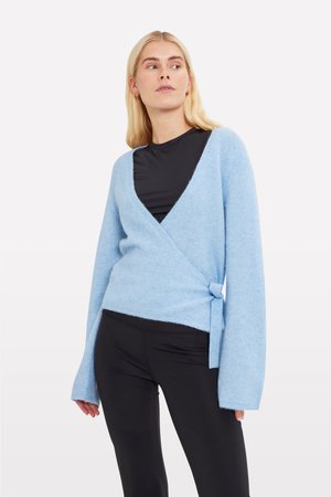 Un pull enveloppant bleu clair en matériau doux, avec des manches longues, un décolleté en V profond et un détail de nœud sur le côté. Porté par-dessus un haut noir.