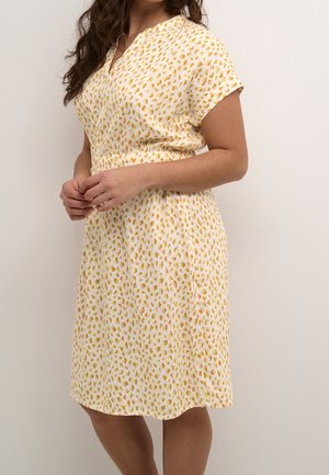 Femme portant une robe blanche à manches courtes et longueur genou, avec des taches abstraites jaunes et orange, debout contre un mur clair et uni.