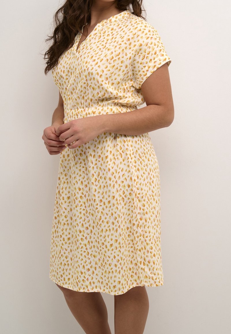 Femme portant une robe blanche à manches courtes et longueur genou, avec des taches abstraites jaunes et orange, debout contre un mur clair et uni.