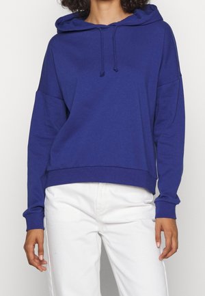 Person wearing a plain blue hoodie with drawstrings and white pants, standing with arms relaxed by their sides.