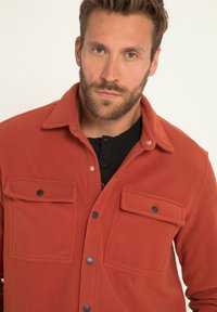 Rostfarbene Fleece-Hemdjacke mit zwei Brusttaschen, schwarzen Druckknöpfen und einem Kragen; getragen über einem schwarzen Hemd mit Knöpfen.