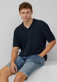 Navy gebreide shirt met korte mouwen en een knoopsluiting, voorzien van een geribde textuur en een klassieke kraag. Gecombineerd met lichtblauwe denim shorts.