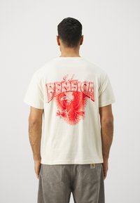 Krēmkrāsas T-krekls ar lielu sarkano ērgli grafiku un uzrakstu "BENFICA" aizmugurē; īsas piedurknes, brīvs piegriezums, gluda tekstūra.