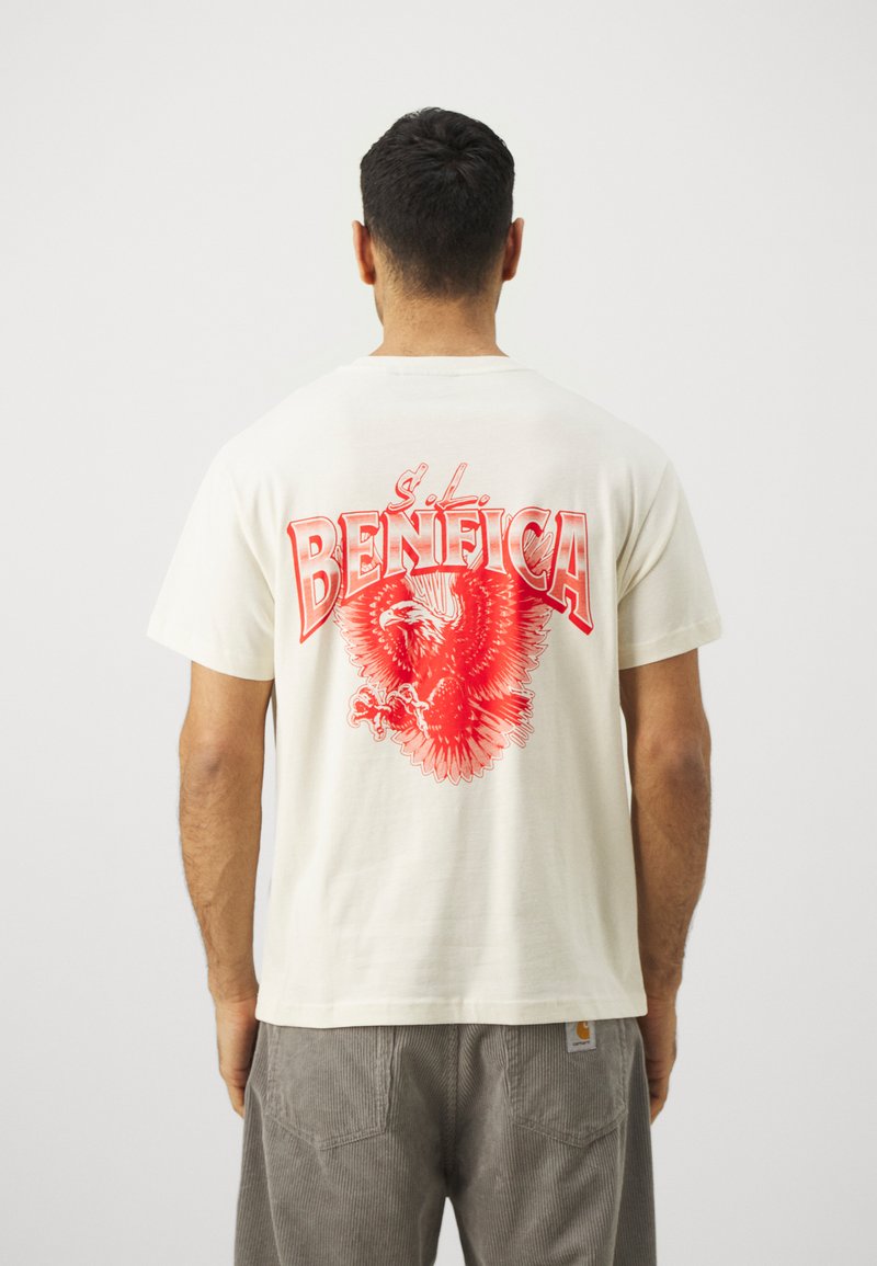 Krēmkrāsas T-krekls ar lielu sarkano ērgli grafiku un uzrakstu "BENFICA" aizmugurē; īsas piedurknes, brīvs piegriezums, gluda tekstūra.