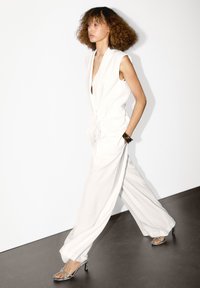 Vit ärmlös jumpsuit med knyt i midjan, vida ben och framfickor. Klara klacksandaler fulländar looken. Slät textur, minimalistisk design.