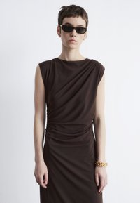 Braunes, drapiertes Kleid mit kurzen Ärmeln und Raffung an der Taille; kombiniert mit kleinen, dunklen Sonnenbrillen und einem goldenen Armband.