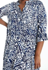 Mörkblå och vit mönstrad klänning med krage, trekvartsärmar och rynkade lager, tillverkad av lättviktig tyg.