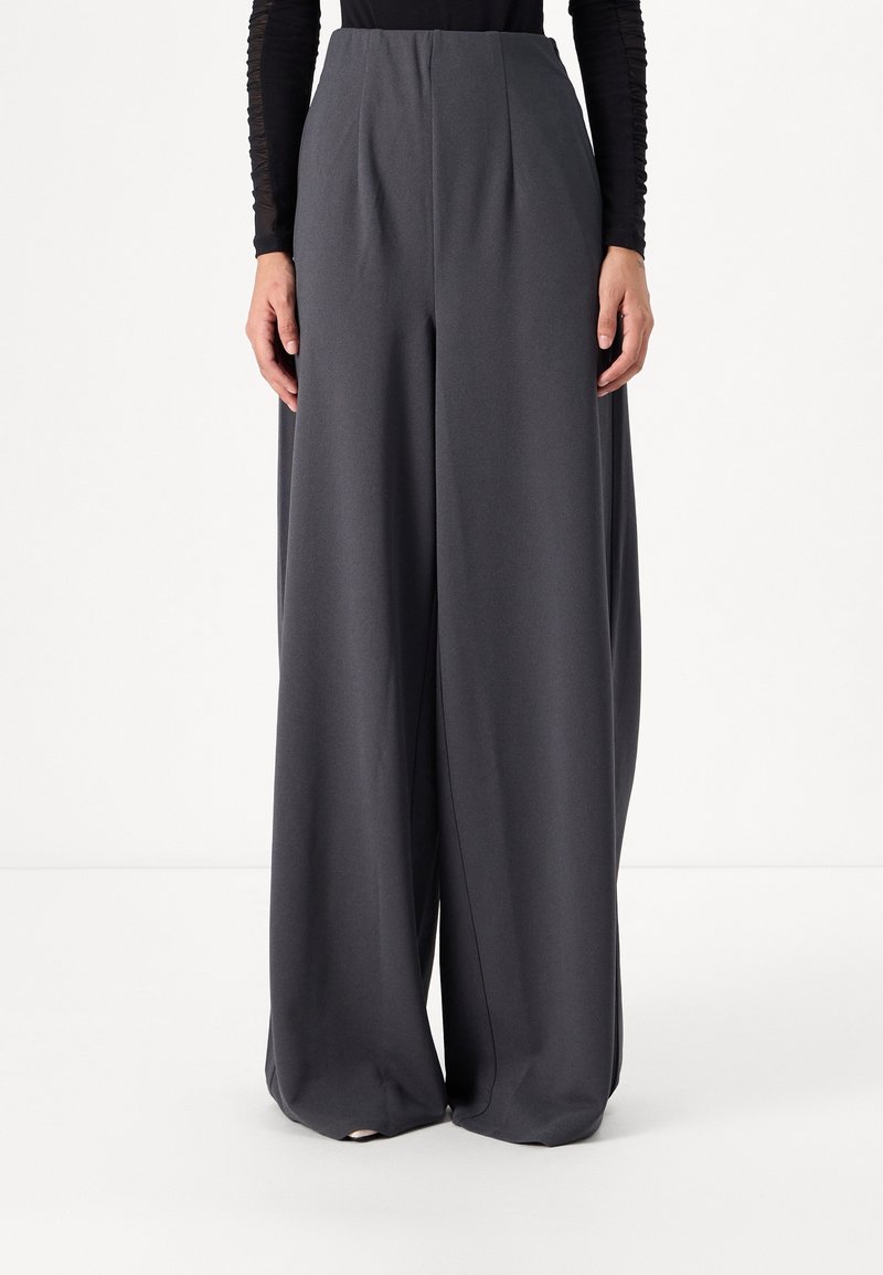 Personne portant un pantalon gris foncé taille haute à jambes larges et un haut noir à manches longues, debout sur fond blanc.