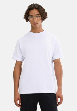 Młody mężczyzna z kręconymi włosami i okularami, ubrany w prostą białą koszulkę i czarne spodnie, stoi na jasnym tle.