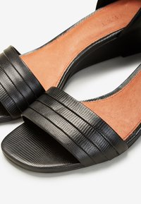 Sandales noires en cuir texturé à bout ouvert avec des larges brides et des semelles intérieures rembourrées marron clair sur fond blanc.