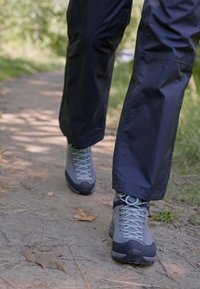 Bottes de randonnée grises avec des lacets gris clair, design mi-mollet, semelles en caoutchouc, portées avec un pantalon imperméable noir, sur un chemin en terre entouré de verdure.