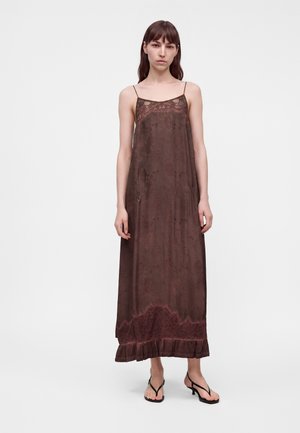 Mujer de pie que lleva un vestido largo marrón con detalles de encaje y tirantes finos, combinado con sandalias negras de tacón bajo sobre un fondo blanco.