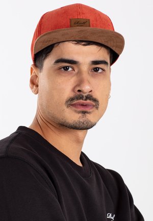 Rote Cordhut mit einem braunen Wildleder-Schirm und einem rechteckigen Patch. Schwarzes Sweatshirt mit Rundhals. Modell mit kurzen dunklen Haaren und Bart.