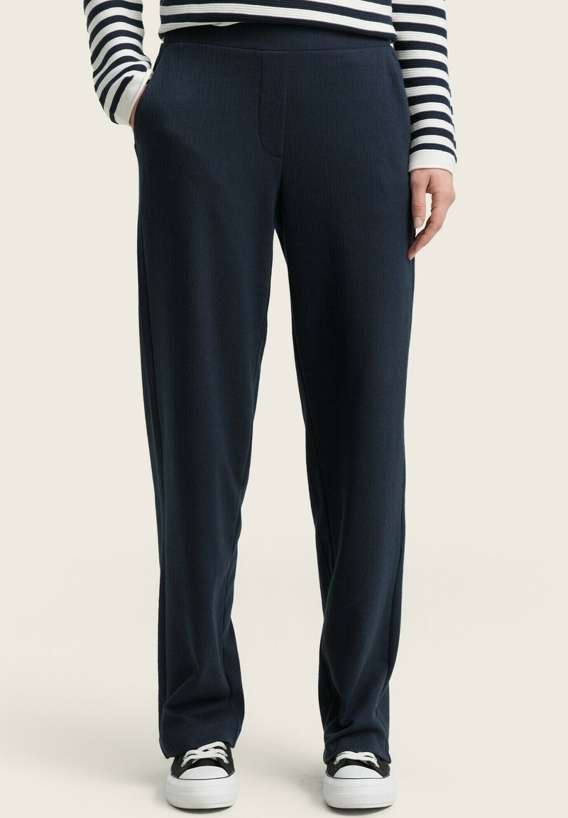 Pantalons bleu marine avec une surface texturée, dotés d'une taille plate, de poches latérales et d'une coupe décontractée, associés à des baskets noires.