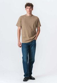 Tan-bearbejdet bomulds t-shirt med korte ærmer, parret med blå denim jeans og sorte sko. Modellen står oprejst mod en ensfarvet baggrund.