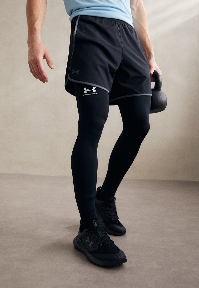 Svarte treningsshorts med reflekterende detaljer over svarte kompresjonsleggings; båret med svarte treningssko. Kettlebell holdt i den ene hånden.