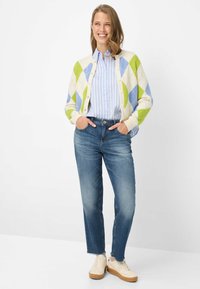 Gebreid vest in crèmekleur met groen en blauw ruitpatroon, gecombineerd met een lichtblauw gestreept overhemd met knopen en medium gewassen spijkerbroek. Beige sneakers.