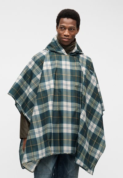 Homme portant un poncho de pluie à carreaux verts, blancs et bleus avec une capuche, par-dessus une chemise marron à manches longues et un jean bleu.